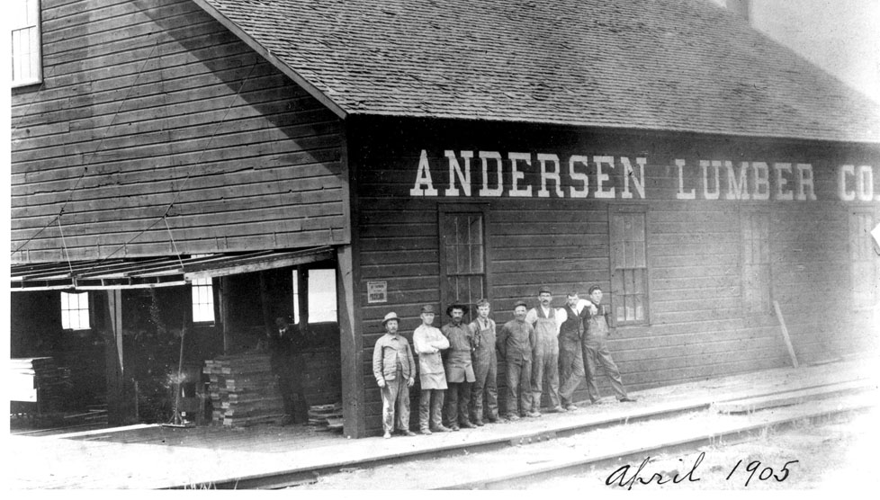 Historic photo from 1903 of eight men outside of a large building labeled "Andersen Lumber"