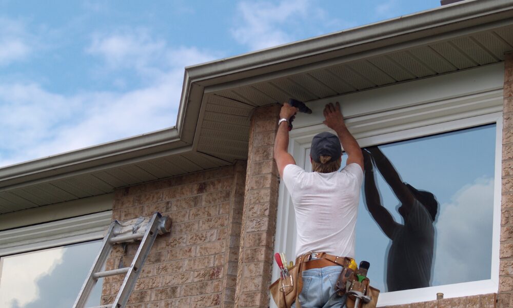 A man on a ladder installs a white picture window on a brick house from the exterior